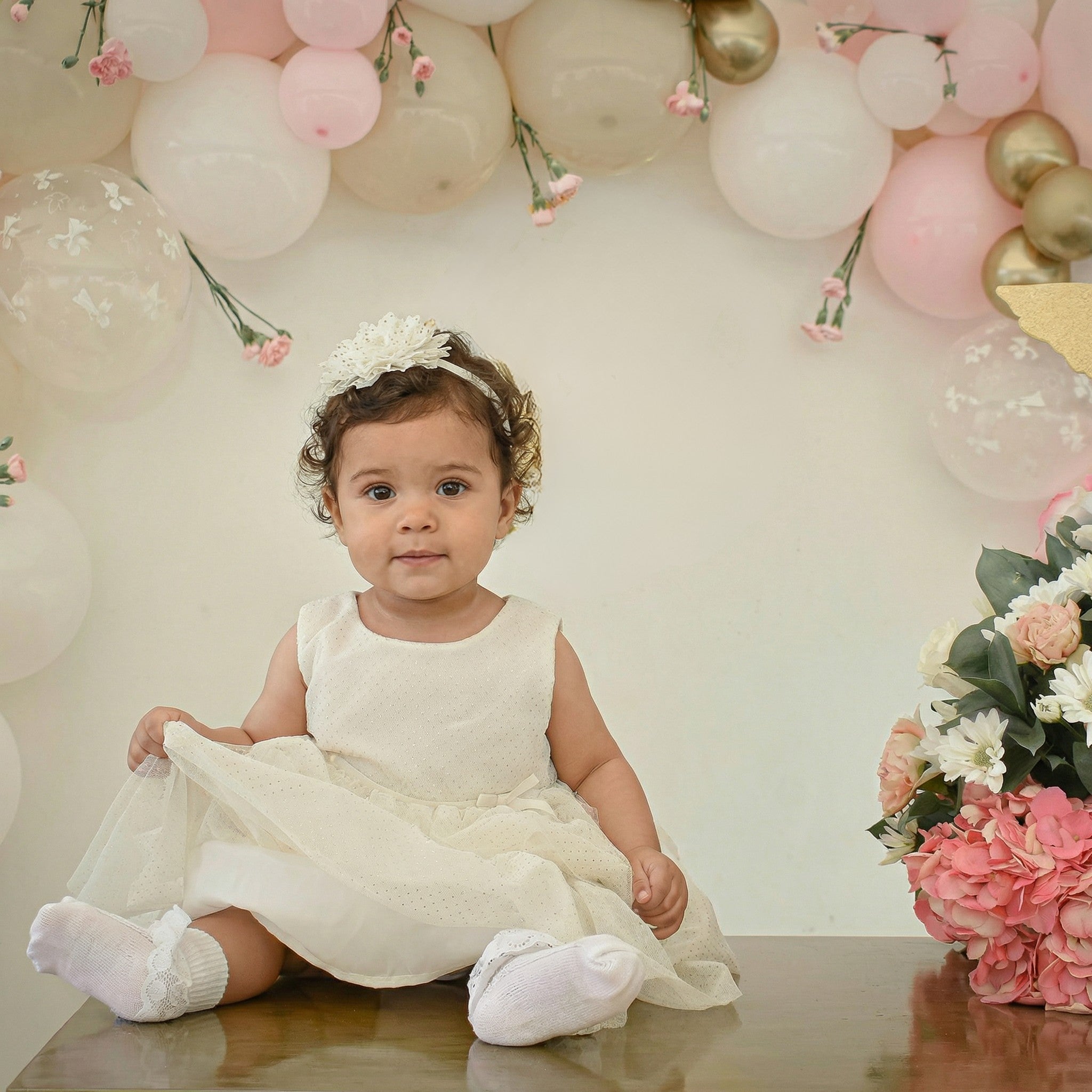 A baby girl wearing an ivory christening gown sitting on top of a table smiling.