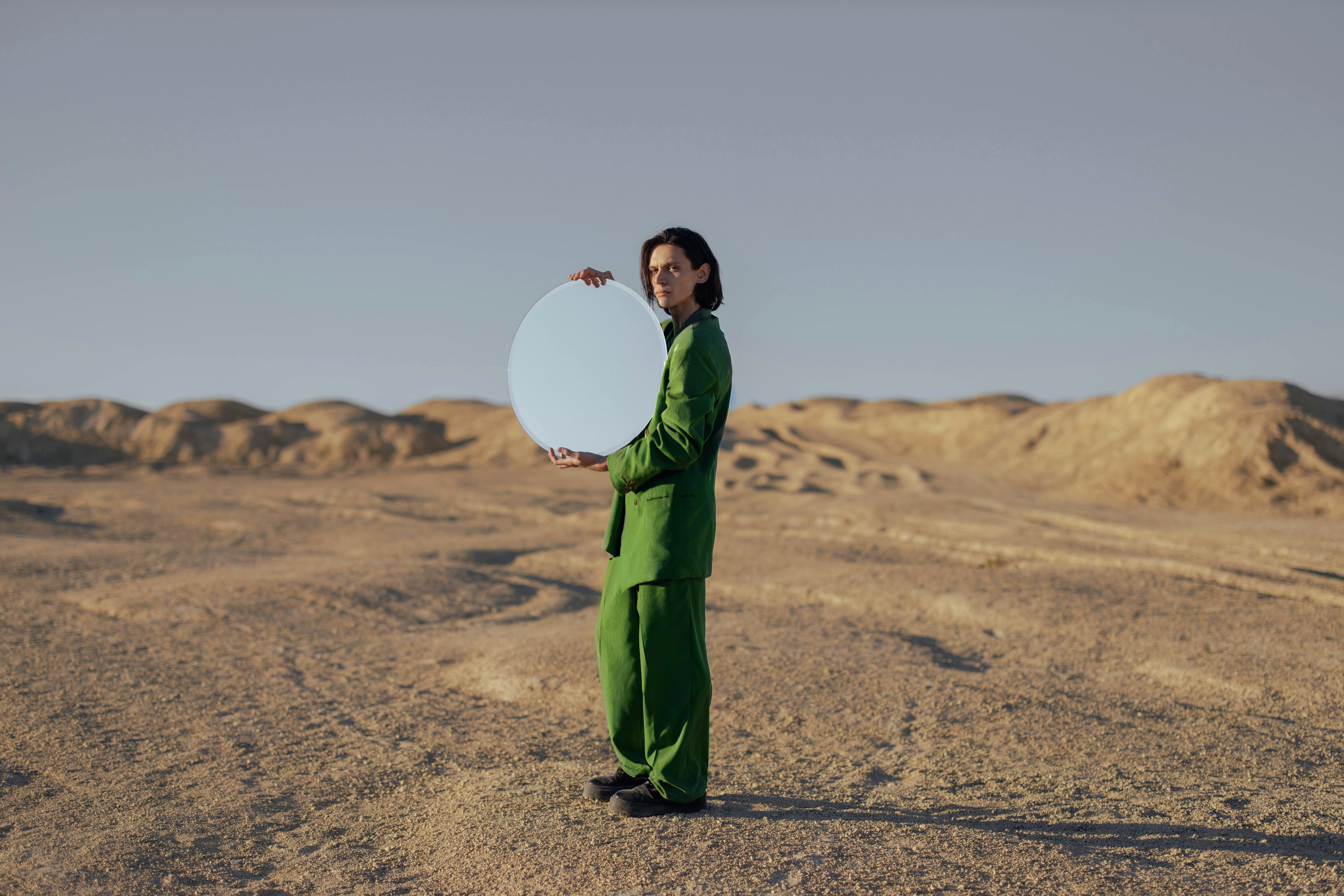 A stylish man in the desert wearing a green tweed suit and holding a circular mirror.