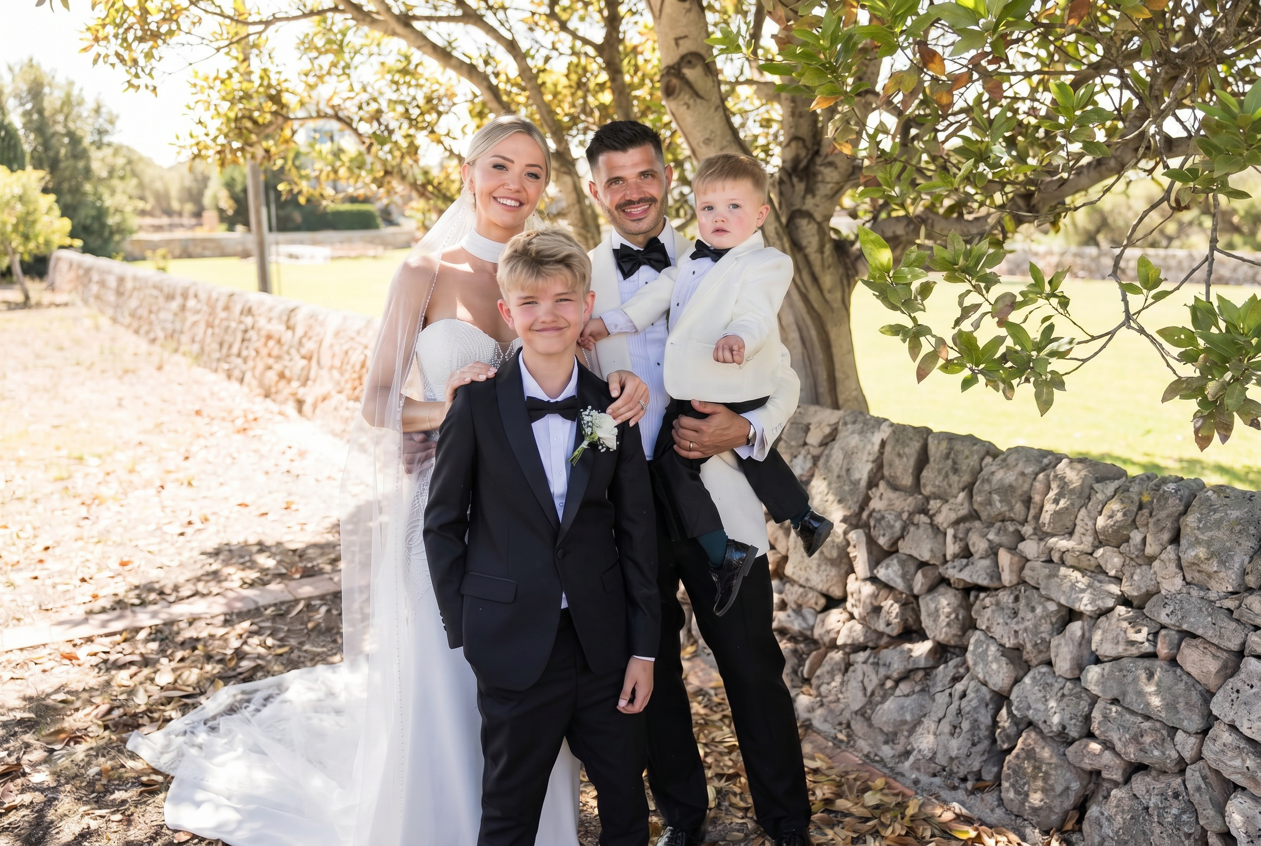 Bride and groom with two page boys wearing black and ivory suits at an outdoor wedding.